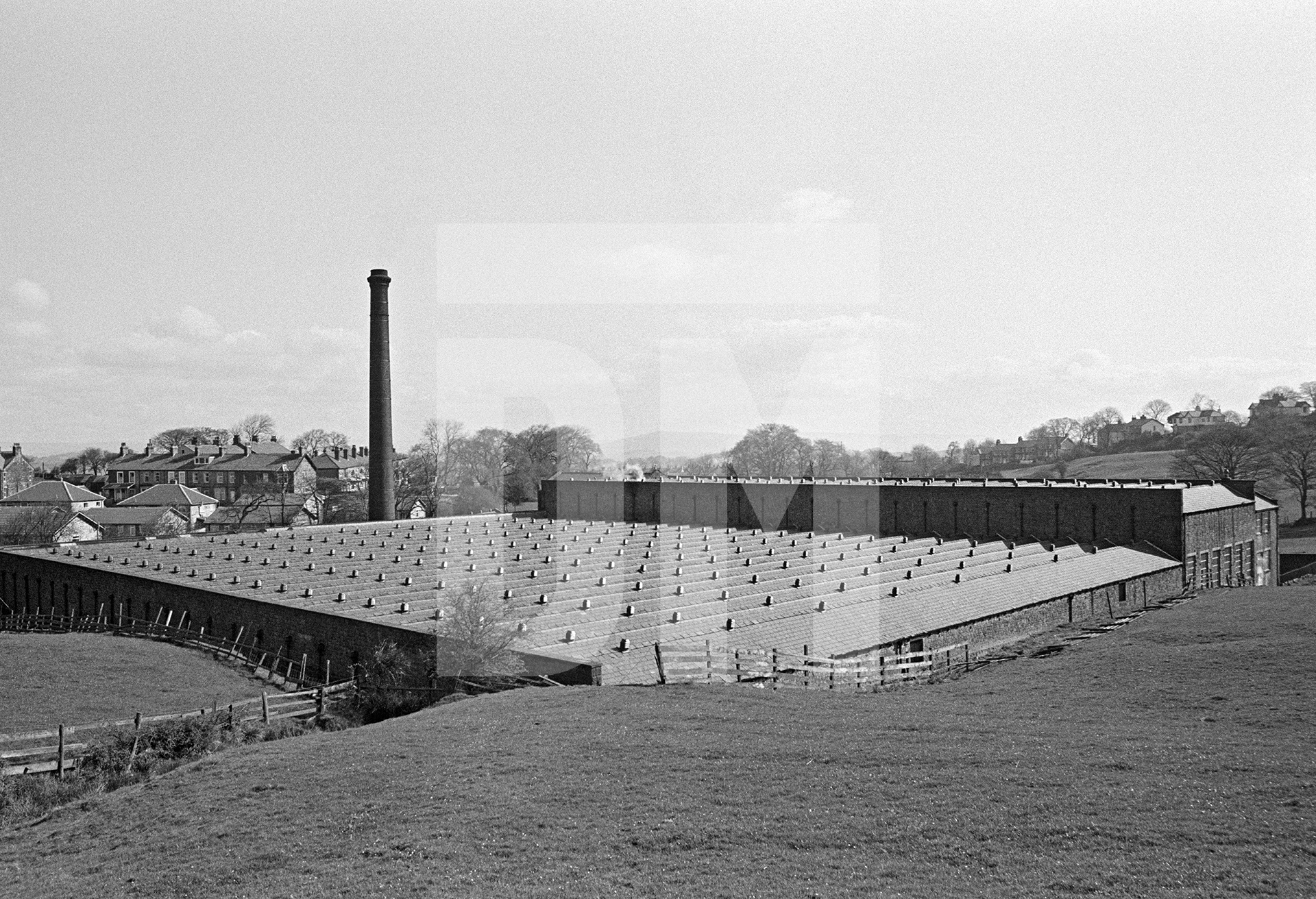 View from the southwest. The slate roof of the weaving shed with the warehouse beyond. The chimney is positioned midway along the northern side. April 1976 by Daniel Meadows