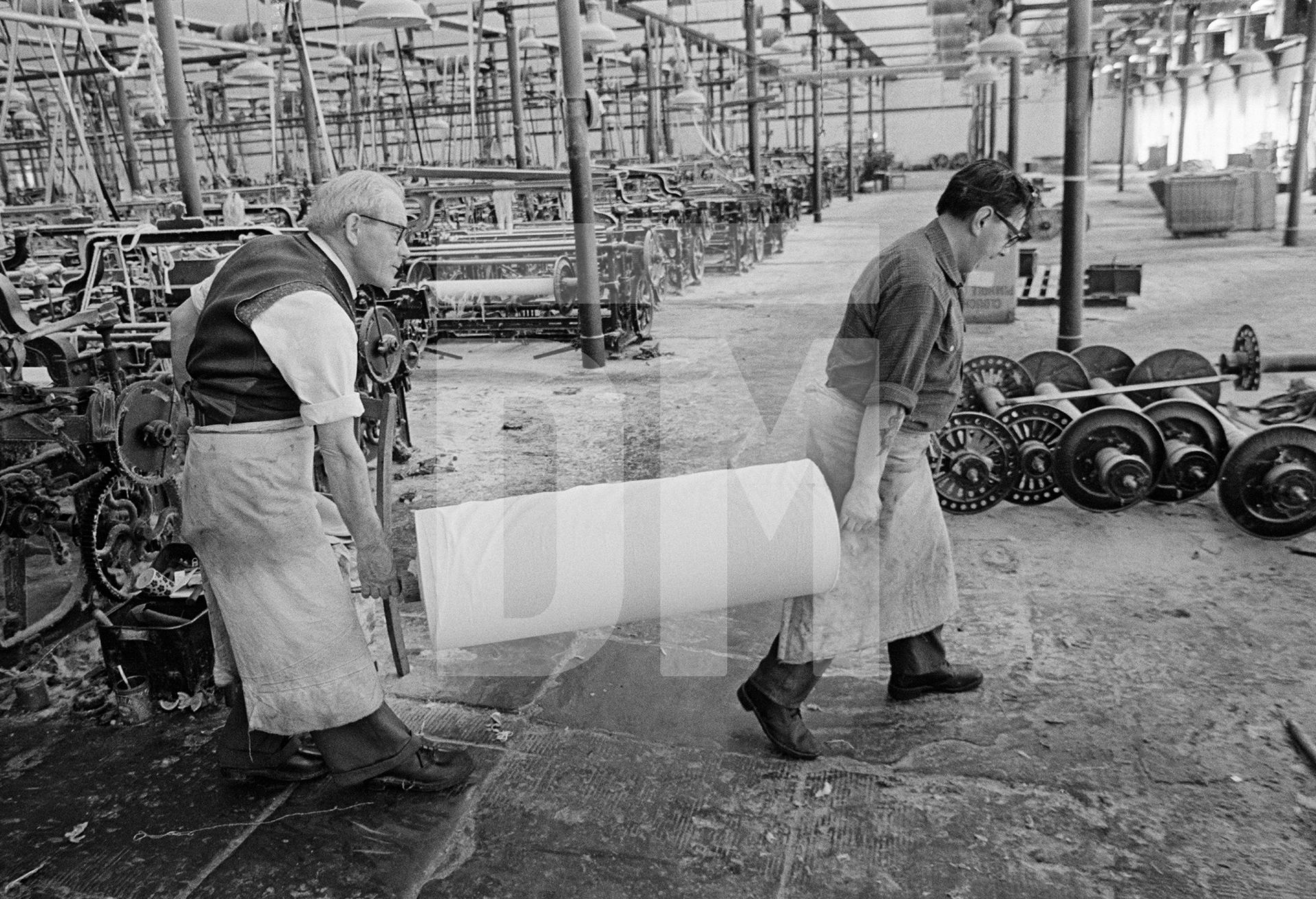 In the weaving shed, cloth carrier and weaver with loom-state cloth. May 1976 by Daniel Meadows