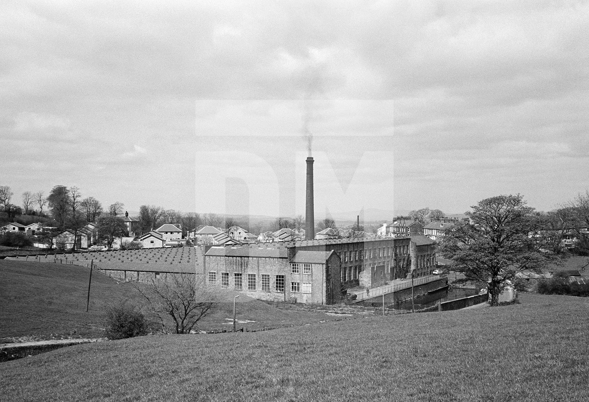 View from the southeast. The office and warehouse face the camera with the ‘dam’ or ‘lodge’ (mill pond) in front of it (to the right in the picture, across the road from the tree). The Engine House is in the northeast corner. In the distance, approx 20 miles north-northeast, is Ingleborough (mountain) in the Yorkshire Dales. April 1976 by Daniel Meadows