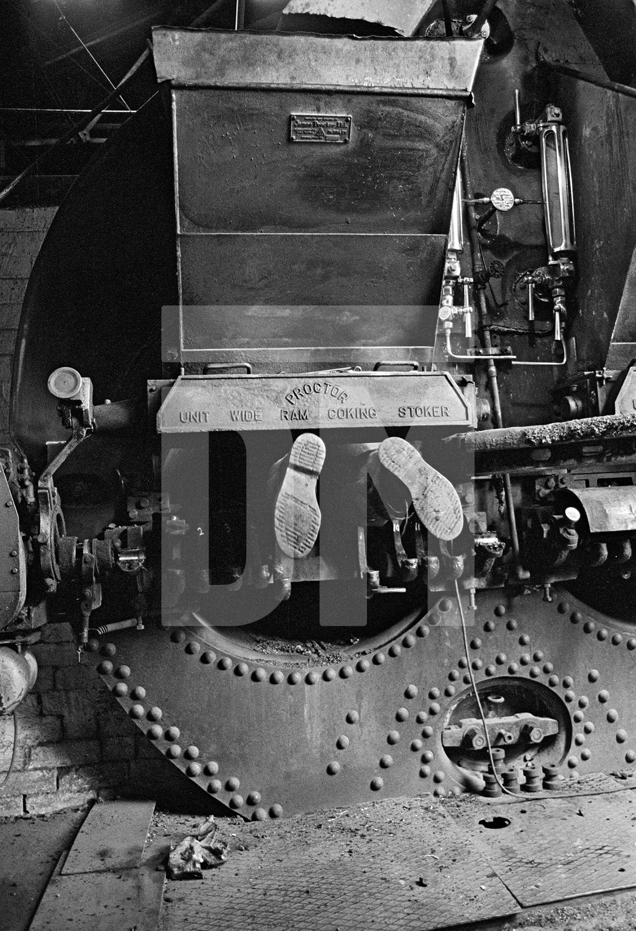 Only the feet of engineer Stanley Graham can be seen as he works on the fire bars in the Lancashire boiler. Wakes holiday, July 1976 by Daniel Meadows