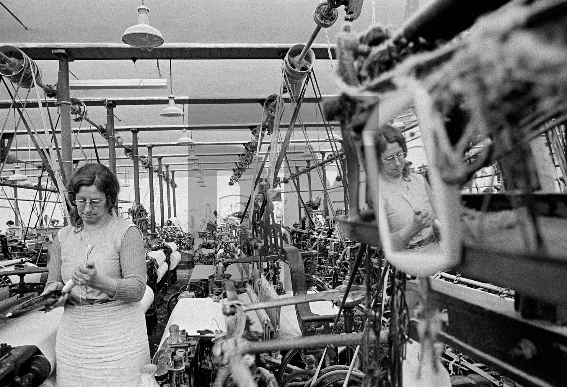A weaver puts a fresh pirn (of weft) into a shuttle. A mirror hangs on one of her looms so that she can comb ‘dawn’ (fluff) from her hair before going home. May 1976 by Daniel Meadows