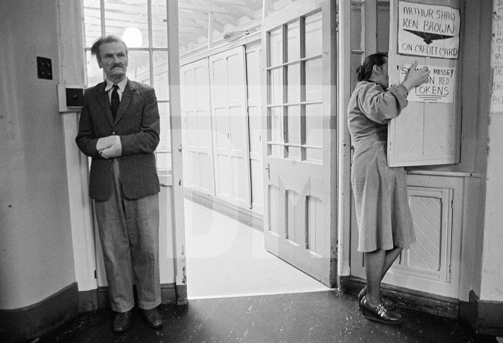 John Keeley, aged 63, stands in the day-room corridor while a nurse searches a cupboard. February 1978 by Daniel Meadows