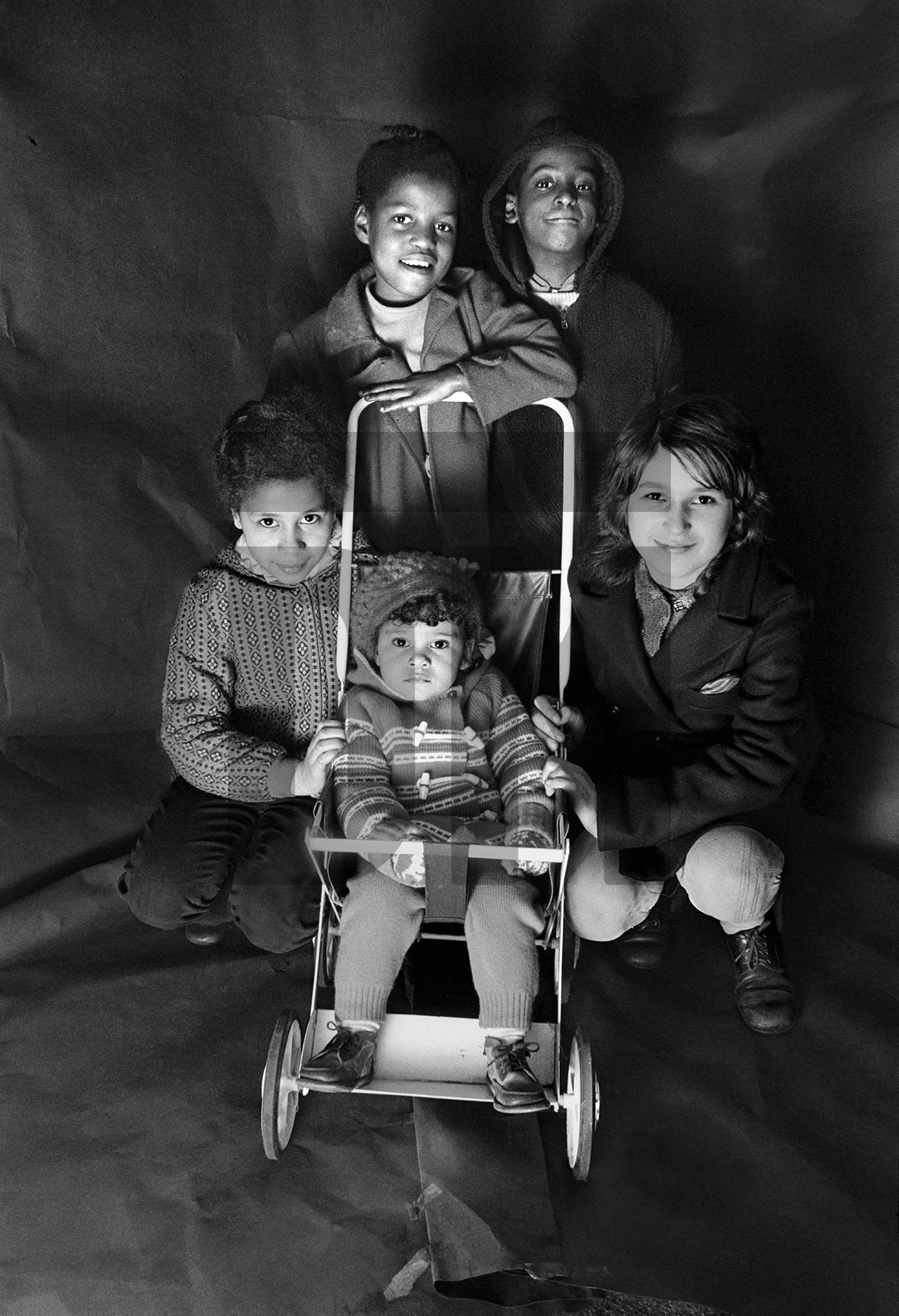 Antoinette Robotham (later Anjee McPherson) and Neville Robotham (later Neville Davis), standing. Debra Caines, crouching left, her sister Tricia in pushchair, Crouching right Wanda Bendix. Group portrait from The Shop on Greame Street, Moss Side, Manchester. February-April 1972 by Daniel Meadows