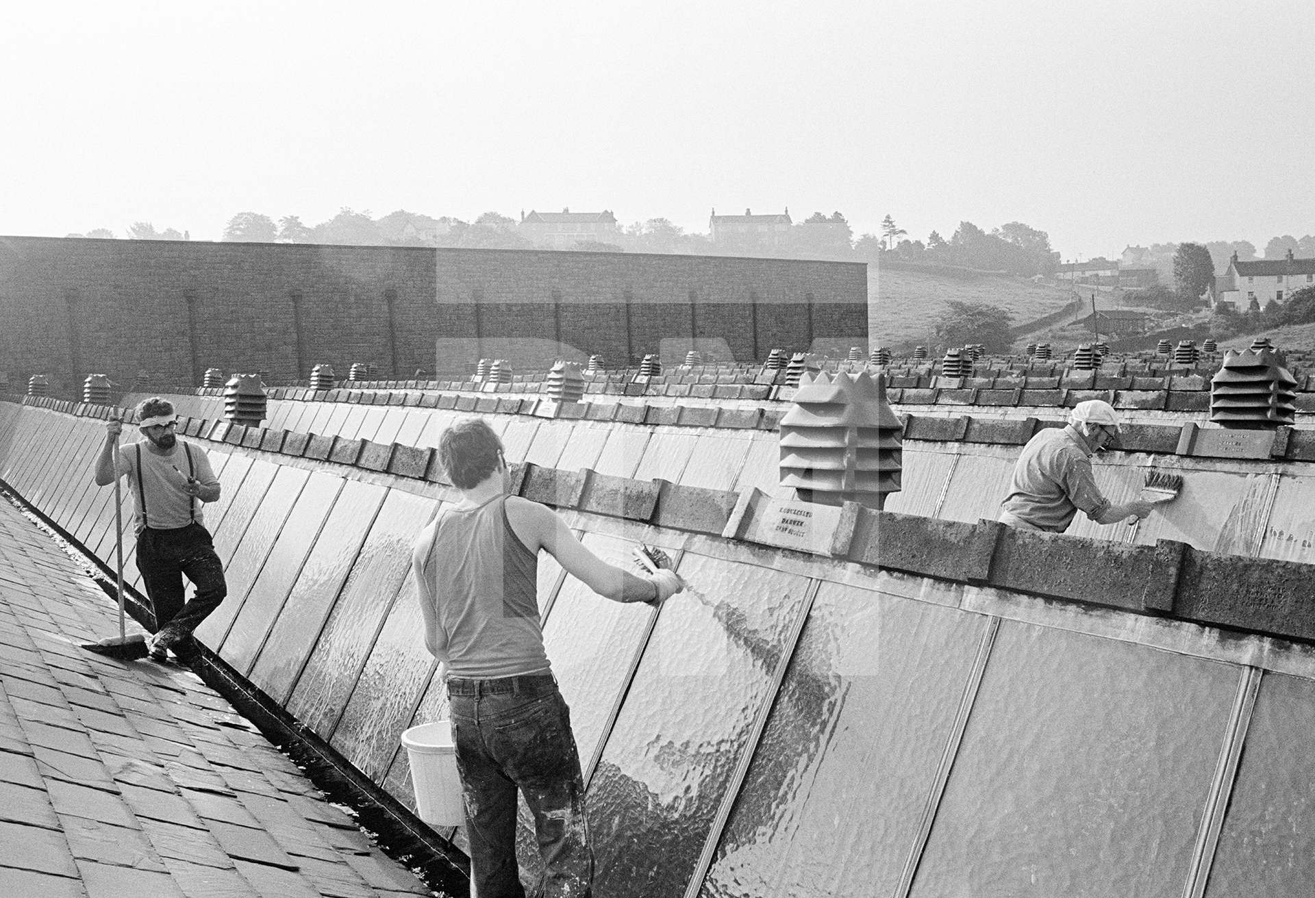Whitewashing the lights (glass) of the shed roof to keep out the summer sunshine and make it cooler for the weavers. Saturday 26 June 1976 by Daniel Meadows