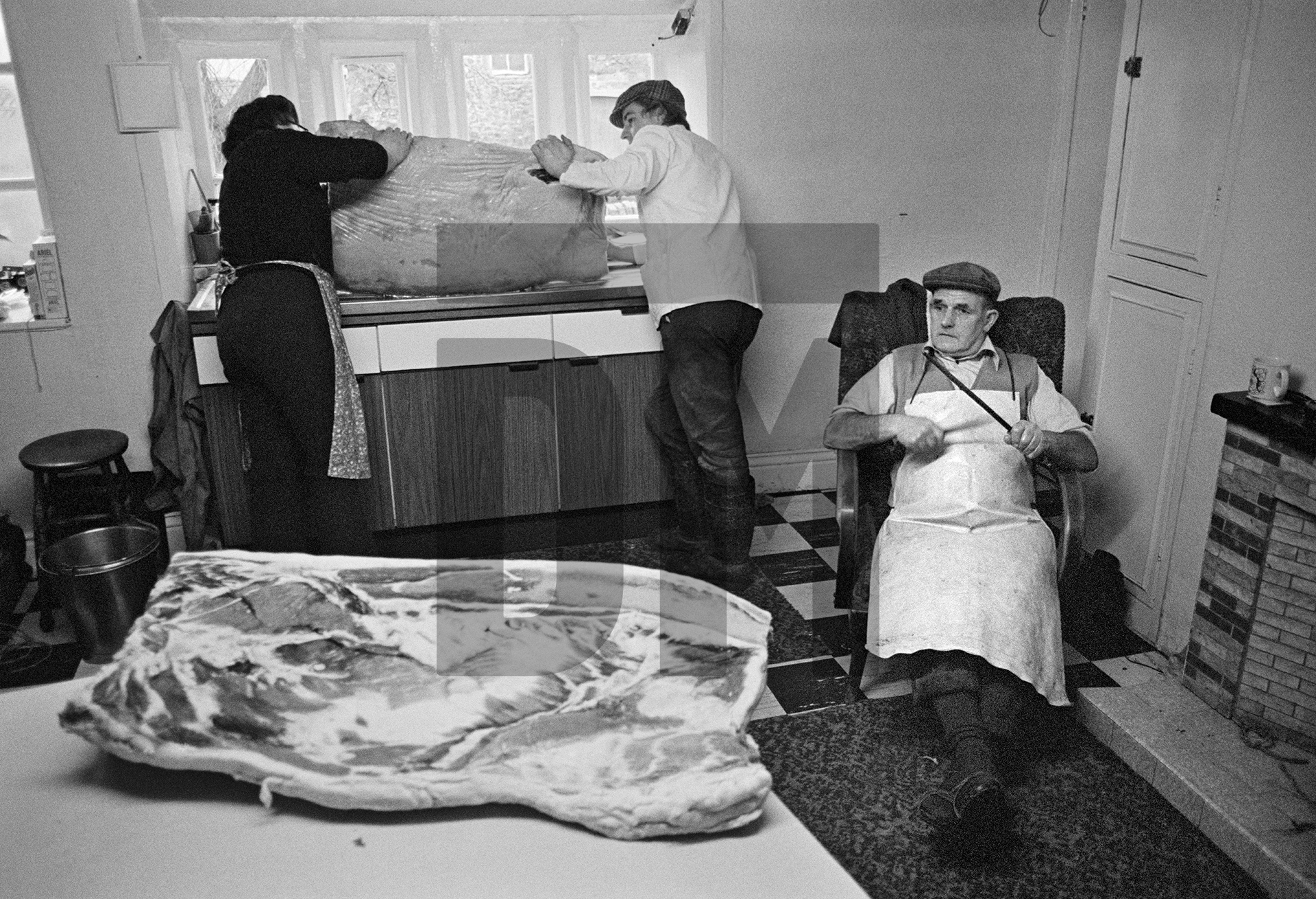 Elsie Richardson and Tony Critchley wash salt off a bacon flitch, Cyril Richardson sharpens his knife. North Yorkshire 1976 by Daniel Meadows