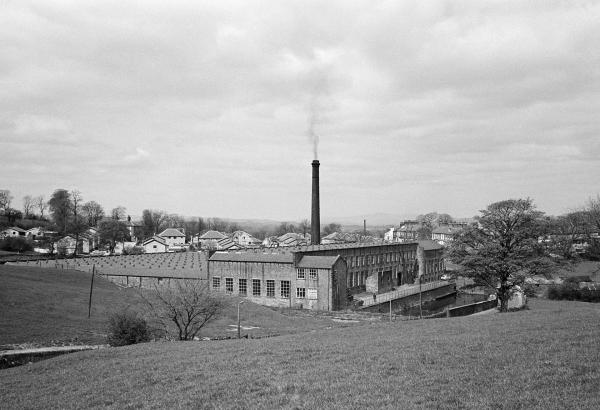 View from the southeast. The office and warehouse face the camera with the ‘dam’ or ‘lodge’ (mill pond) in front of it (to the right in the picture, across the road from the tree). The Engine House is in the northeast corner. In the distance, approx 20 miles north-northeast, is Ingleborough (mountain) in the Yorkshire Dales. April 1976