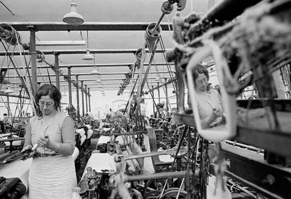 A weaver puts a fresh pirn (of weft) into a shuttle. A mirror hangs on one of her looms so that she can comb ‘dawn’ (fluff) from her hair before going home. May 1976