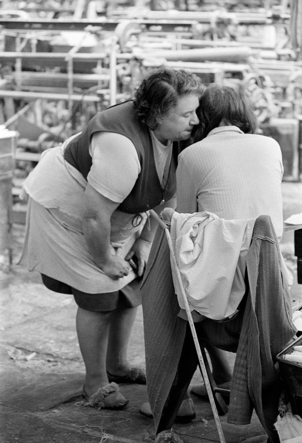 In the weaving shed, where the roar of the looms is deafening, two weavers have a private conversation, their mouths concealed from the eyes of their lip-reading workmates. April 1976